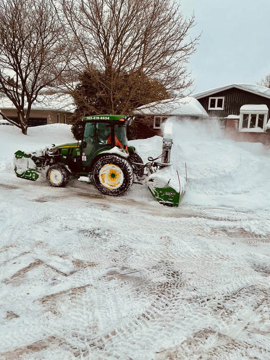 Green snow tractor using a snow blower to clear snow from a residential driveway.